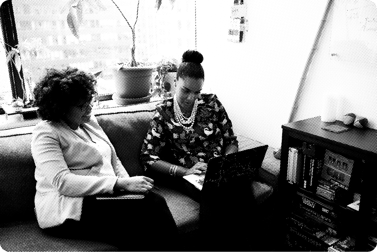 Two women look at a laptop while sitting on a couch. 