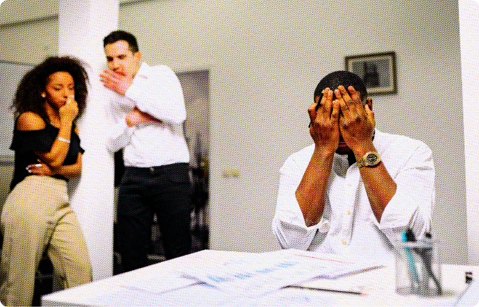 Man sits at a table frustrated with his hands covering his face while two coworkers stand next to him